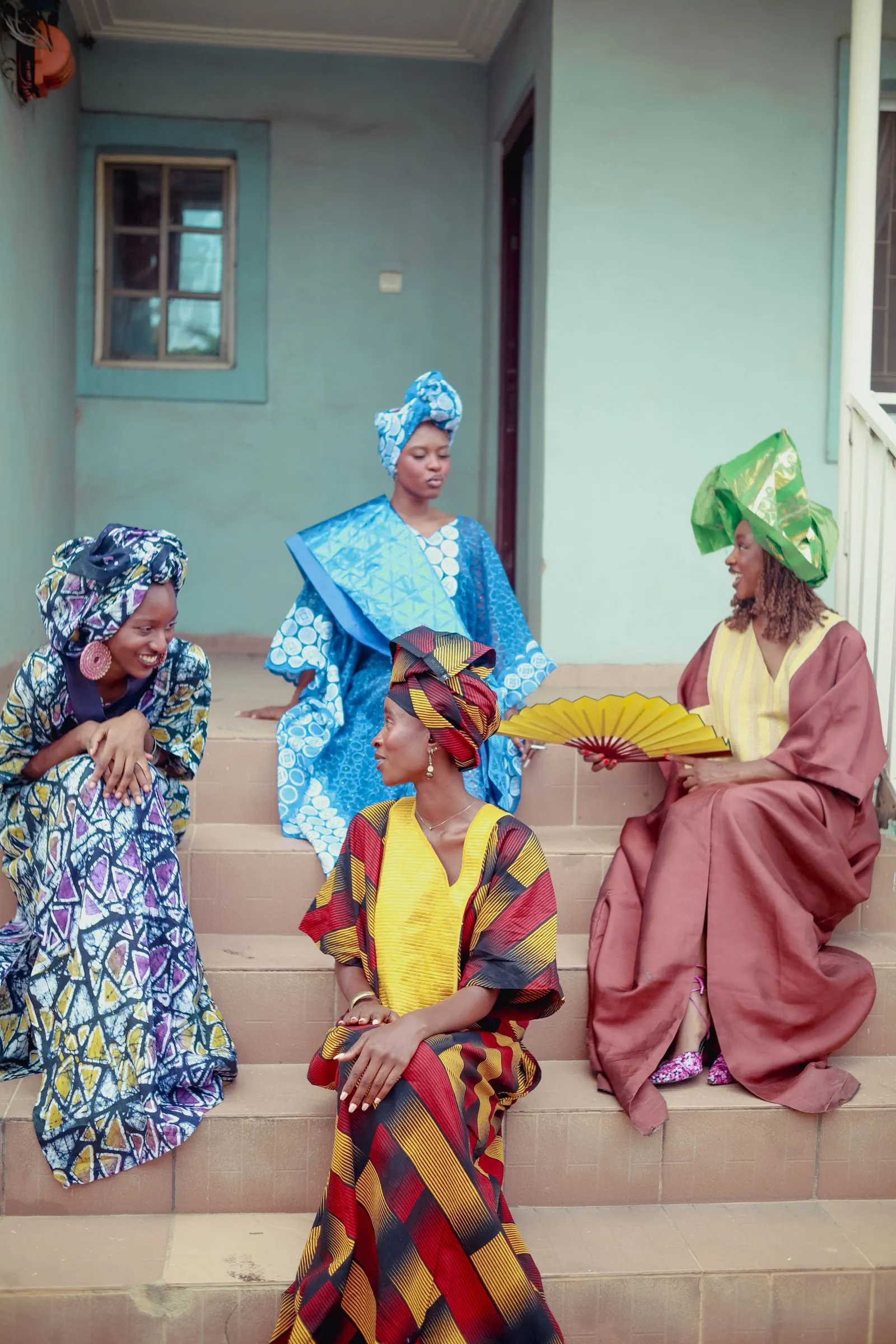 Four women in bright wrappers and geles talk together on a set of steps